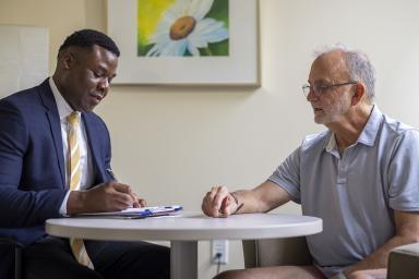 Older man sitting at table with doctor in blue blazer discussing report on clipboard. 