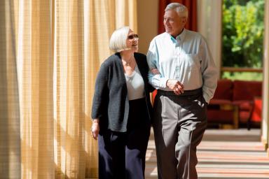 An older man and woman walk down a sunlit corridor.