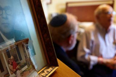 Older man wearing a Kippah sitting near some photos with another older man.