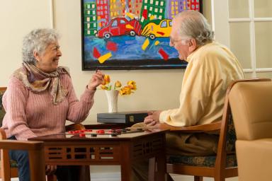 Older man and woman sitting at a table in a lounge area playing checkers.