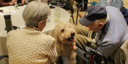 An older male and female sit at a table and look down at a white dog