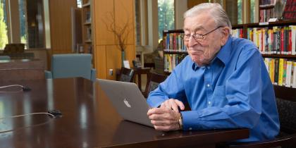 An elder male sits and smiles at a laptop while sitting in a library