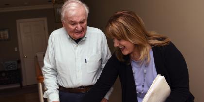 A Home Health worker smiles while helping a male patient with his walker