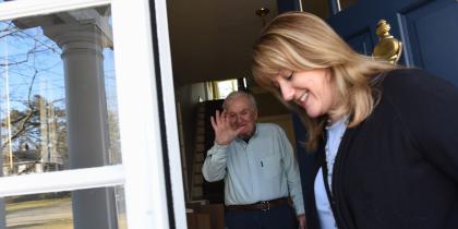 A Home Health worker smiles while standing by a door while a male patient pushes a wheelchair towards her