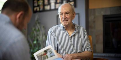 Older man sitting while another man is looing at a photo book
