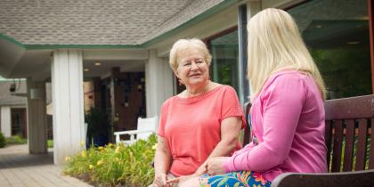 Older woman sitting on a bench outside talking with another woman.