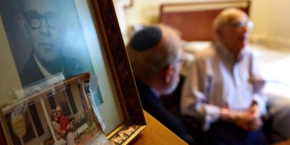 Older man wearing a Kippah sitting near some photos with another older man.