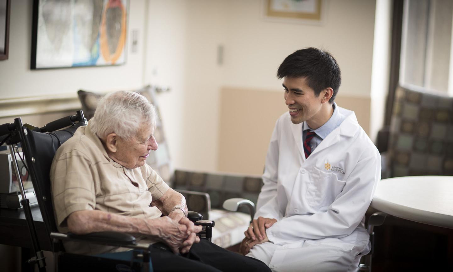 An older man in a wheelchair sits talking with a young Harvard Medical School doctor at Hebrew SeniorLife. The doctor is smiling and wearing a white coat.