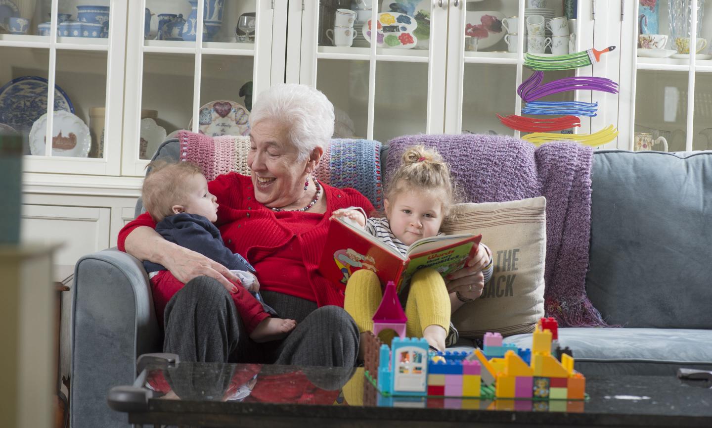 Grandmother in red sweater sits on couch with two grandchildren, a baby under one arm and a toddler with a book under the other. Brightly-colored blocks sit on a coffee table.