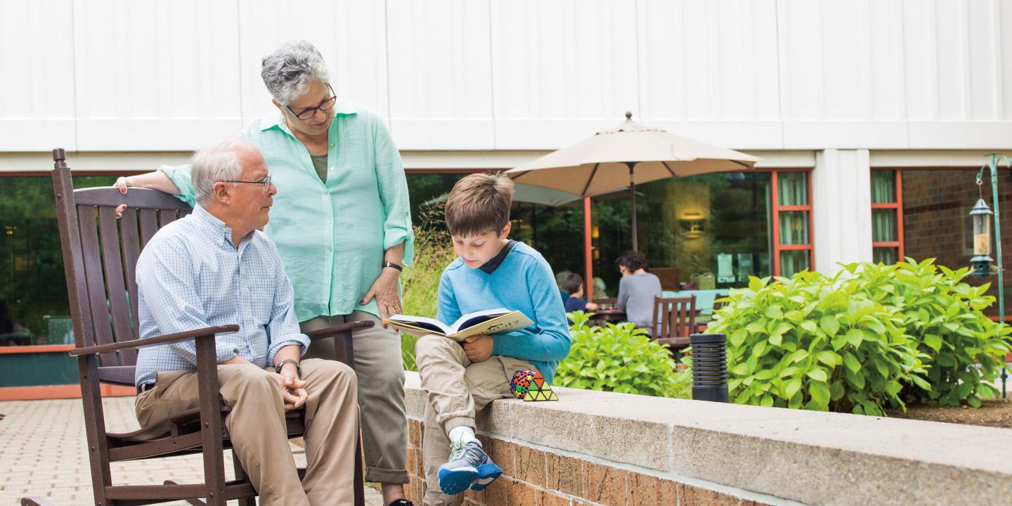 Grandparents spend time reading with a grandchild in the courtyard of a Hebrew SeniorLife senior living community.