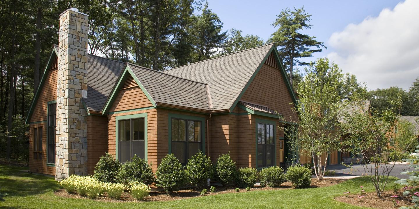 A cottage residence at NewBridge on the Charles sits on a corner lot with lush landscaping, large windows, a stone chimney, wooden façade, and two-car garage. 