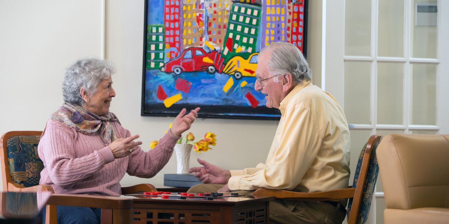 An older man and woman at Center Communities of Brookline play a game of checkers, near a brightly colored painting of city life