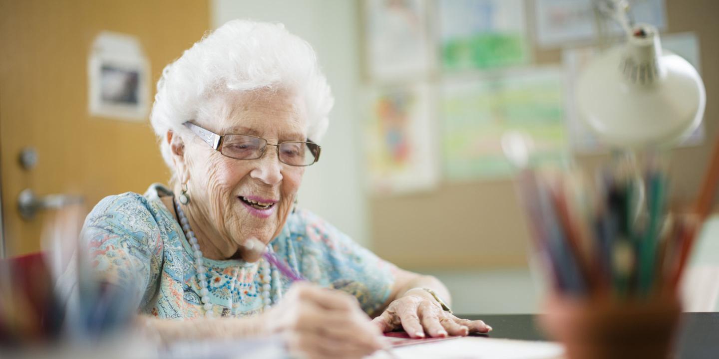 An older woman in a bright floral top holds a pencil and draws on a piece of paper.