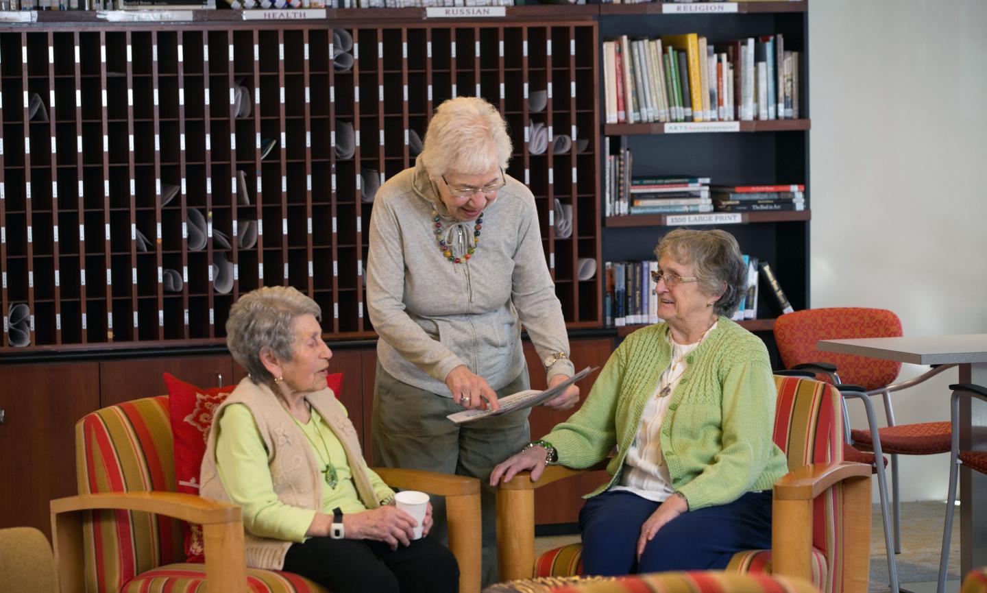 Three women drink coffee and converse in a library seating area at Center Communities of Brookline