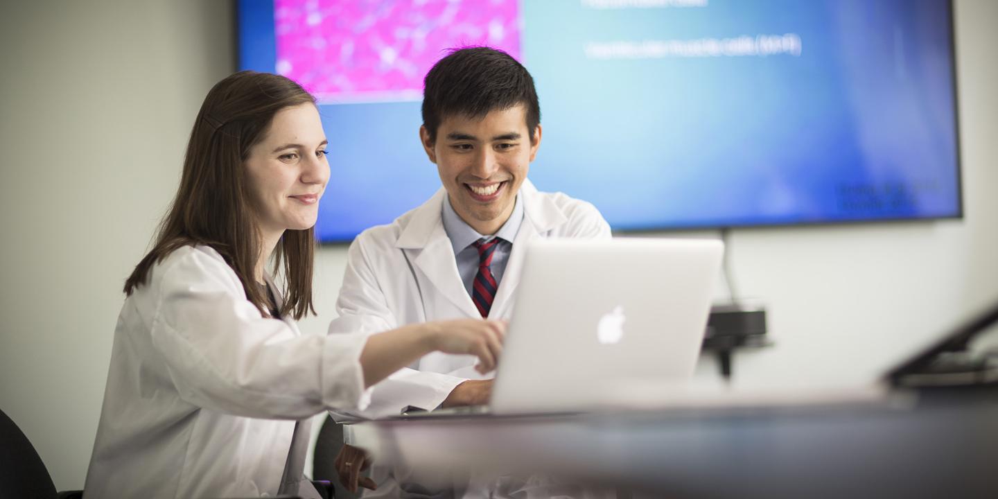 A female and a male Harvard Medical School fellow sit at a table at Hebrew SeniorLife looking at a laptop. They are wearing white coats.