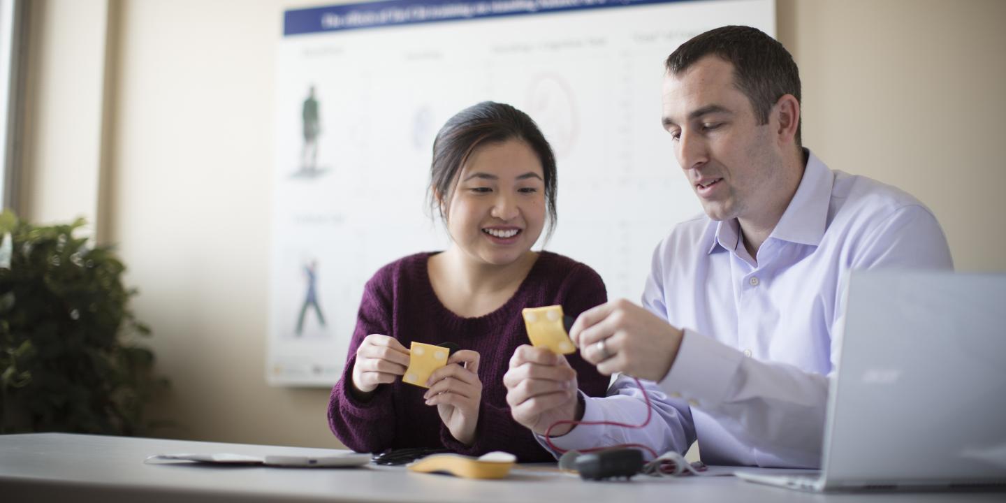 A female and a male researcher from the Hinda and Arthur Marcus Institute for Aging Research sit at a table with a laptop. They are each holding a small yellow square that has wires attached.