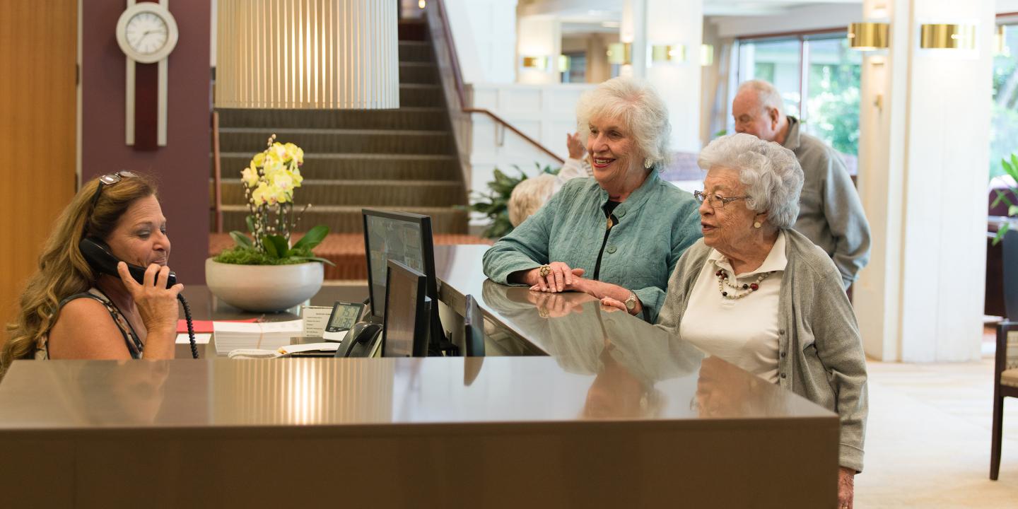 Two residences talk to front desk receptionist who is on the phone to get them what they need.