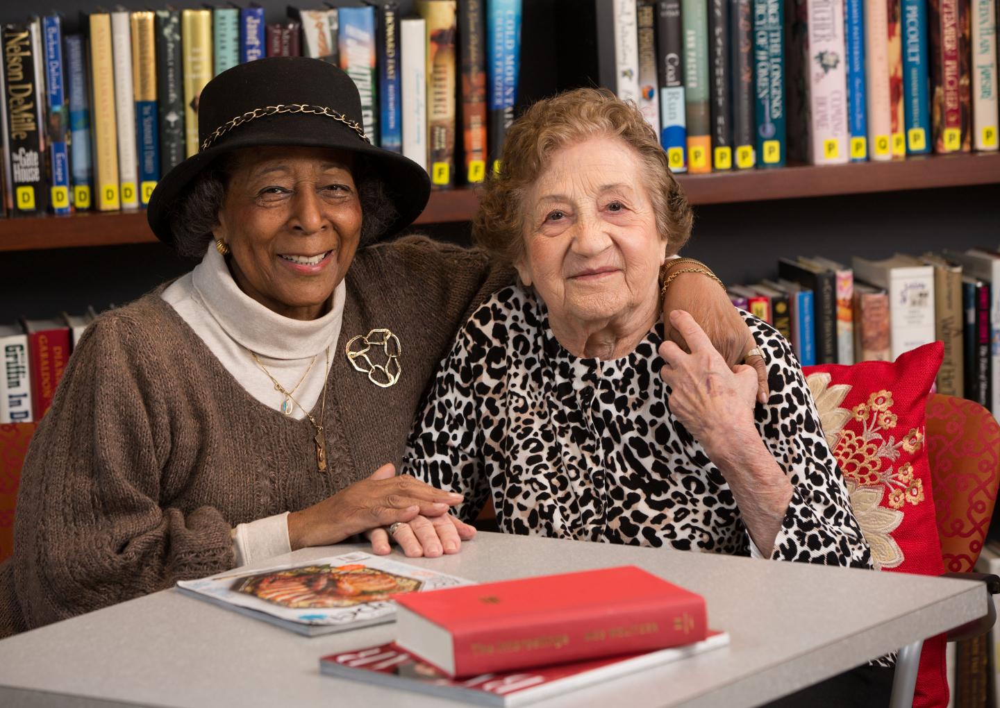 Two older women sit together in a library at a table with books and magazines.