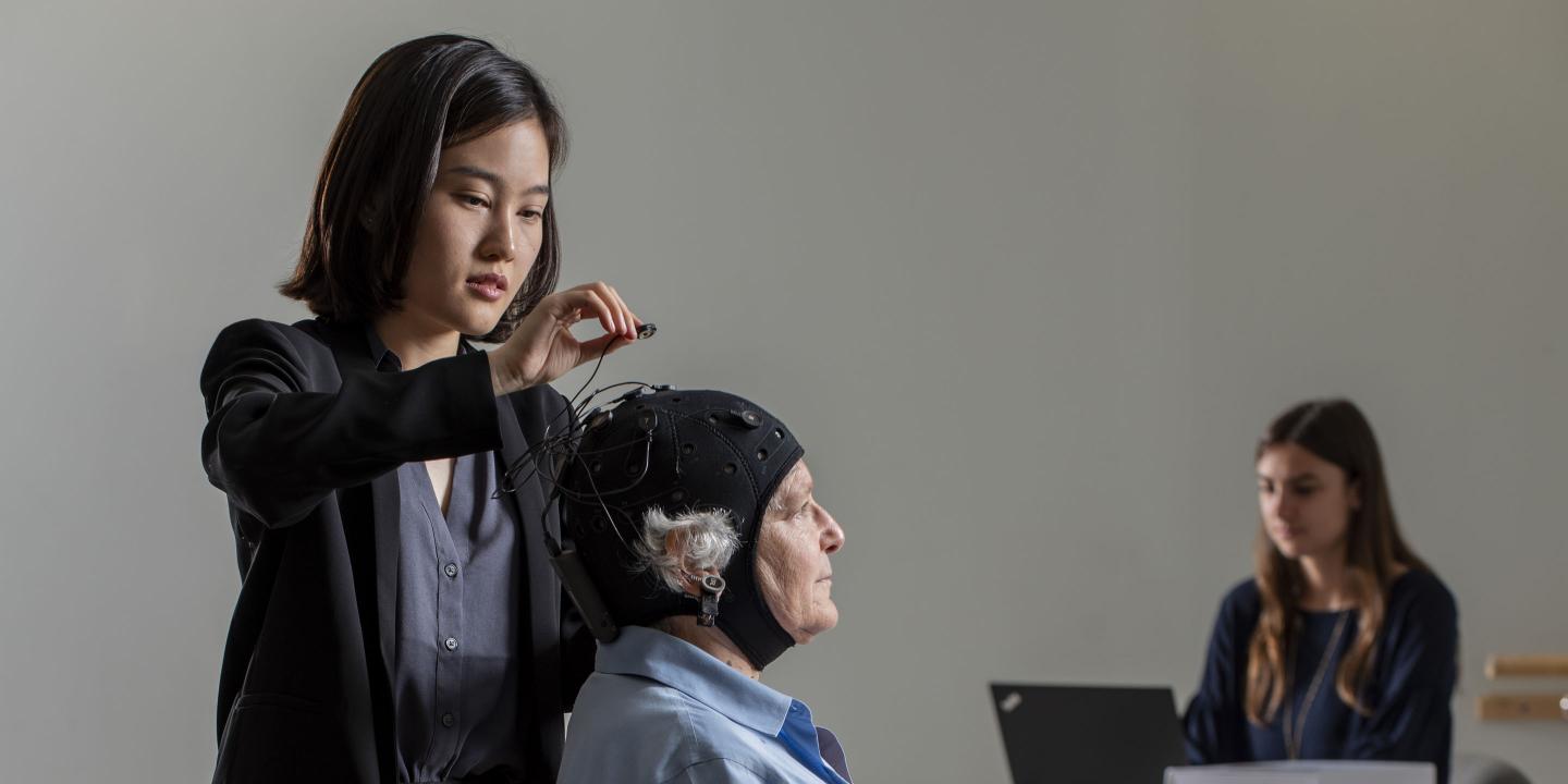 An older woman sits in a chair while a younger woman attaches wires to a black brain stimulation cap the older woman is wearing