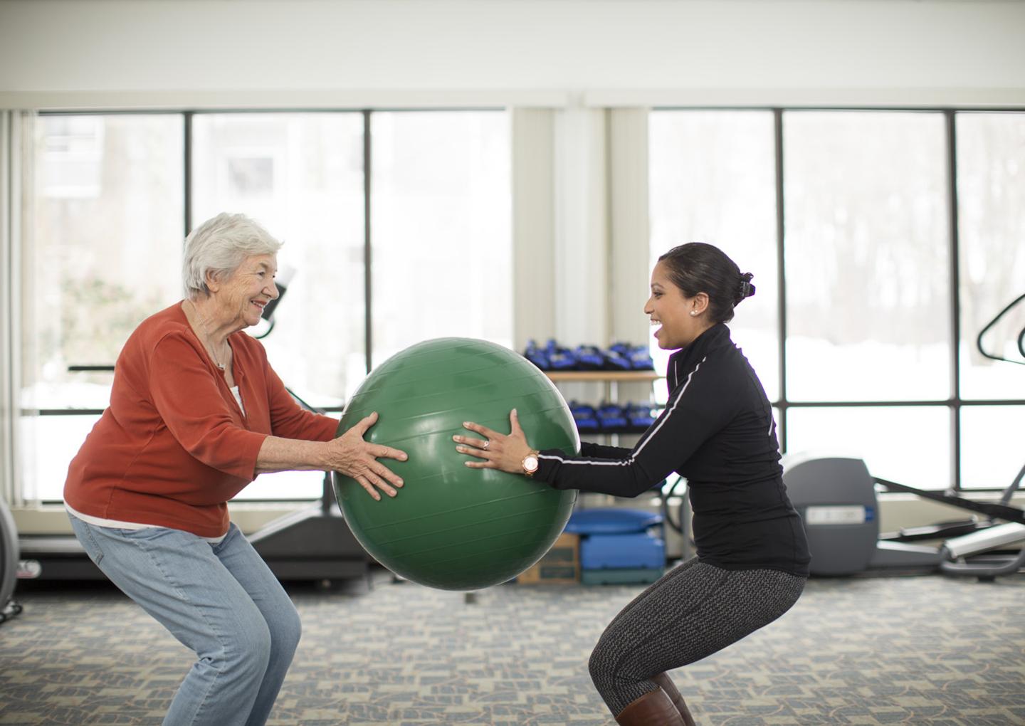 An older woman on the left and younger woman on the right stand facing each other, standing in a squat position, holding a large exercise ball between them. They are in a gym with large windows and exercise equipment in the background.
