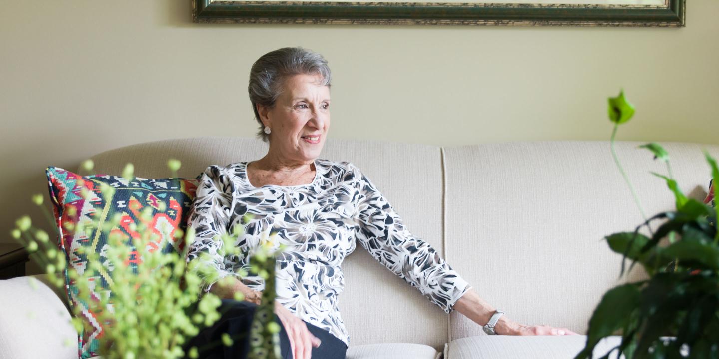 A stylish older woman in a black and white patterned shirt sits comfortably on her white sofa with the sun shining through a nearby window and with plants in the foreground.