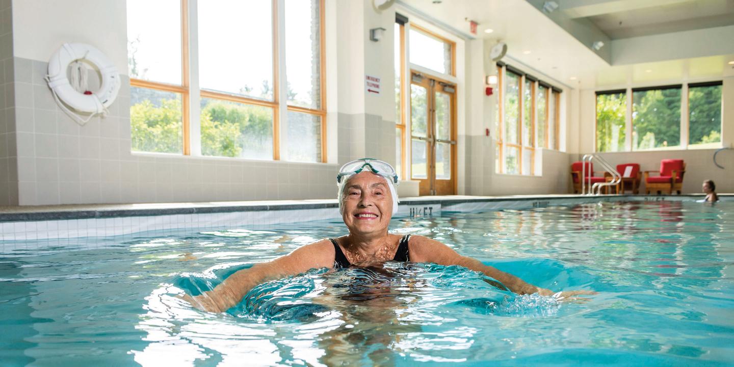 An older woman smiling and swimming in a large sunny indoor pool surrounded by windows.