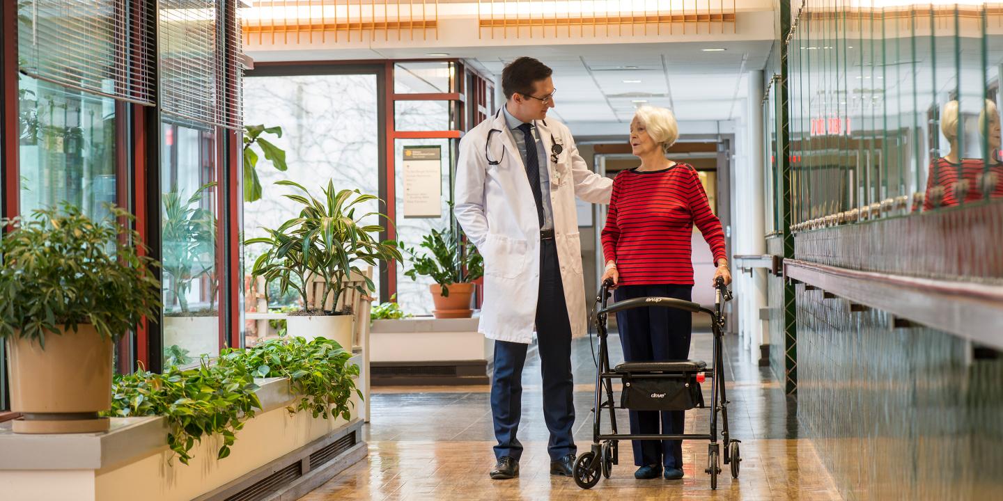 A gray-haired woman with a rollator stands talking to a male doctor in a hallway at Hebrew Rehabilitation Center in Boston.
