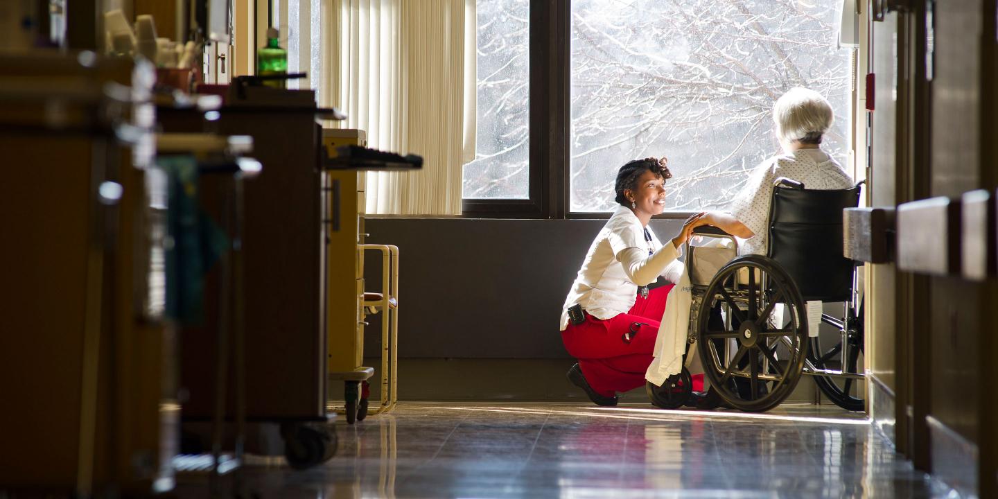 A nurse visits with an older woman in a wheelchair in front of a large picture window.