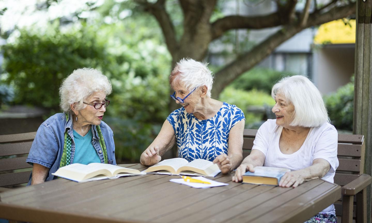 Three older women sit at a table outside reading books