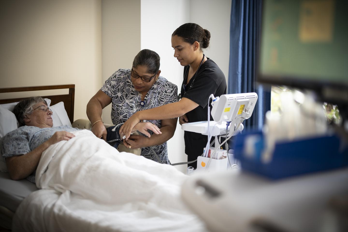 A nurse shows a certified nurse assistant how to use a blood pressure cuff on a patient.