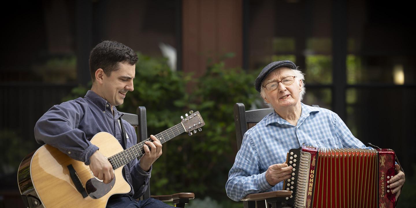 A music therapist plays his guitar while an older gentleman plays his accordion.