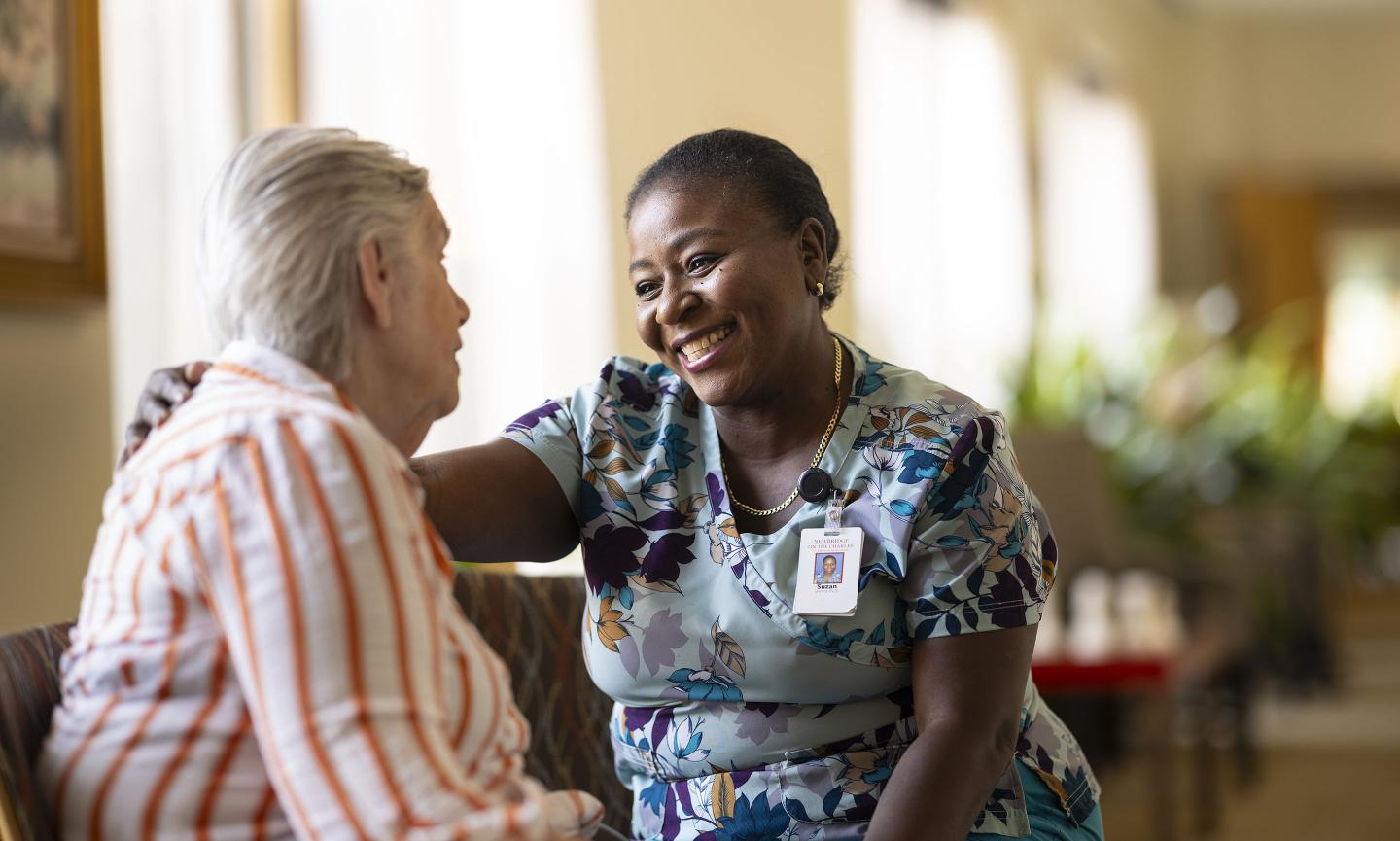 A patient care assistant smiles while speaking with an older female patient.