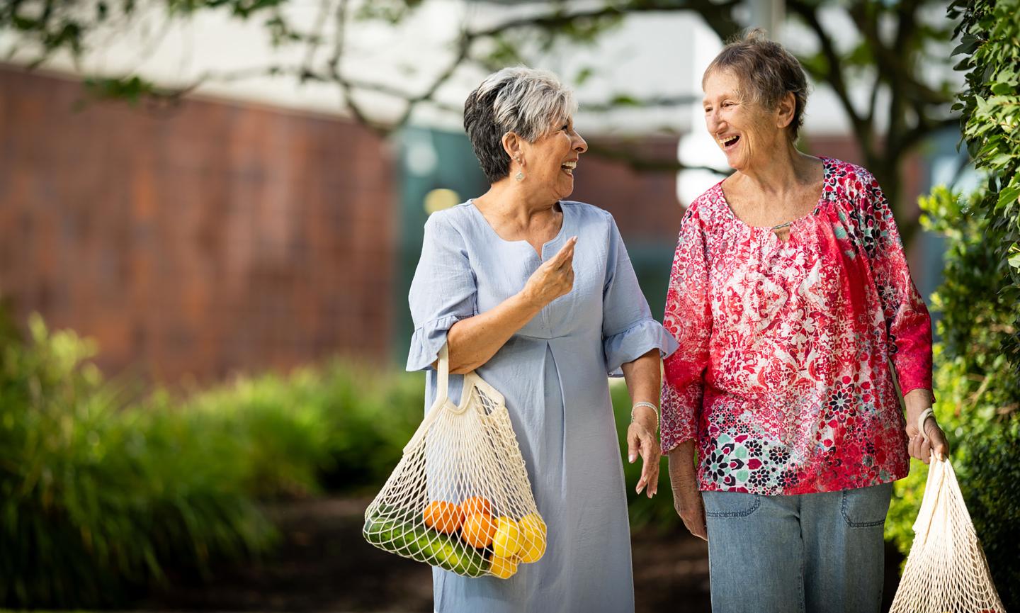 Two older women walk down a path with bags of produce.