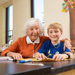 Older woman sits with her arm around a young boy as they work on an art project together.