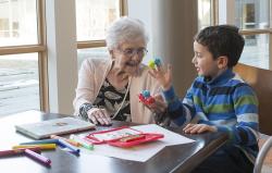 Older woman sits at a table with a young boy as they play with colorful finger puppets.