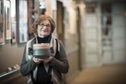 An older woman in gray sweater standing in a hallway gallery at Orchard Cove holds a ceramic pot she has made.