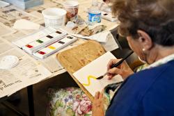 An older woman in a blue shirt and brightly flowered apron paints a yellow brush stroke as she begins a watercolor in an art workshop.