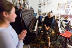 Young girl stands in foreground as an older woman sits at a piano in her apartment as part of a program to bring visitors to seniors in their homes.