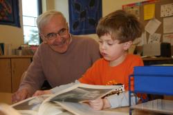 An older man and young boy sitting next to each other at a table. The boy is looking at a book and the older man is looking on and smiling.