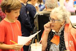 An older female sits and looks intently at a school-age boy who stands in front of her, reading from a piece of paper.