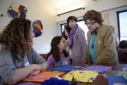 A woman and her younger daughter sit at a table that is covered with multicolor construction paper and craft supplies. They are smiling at an older woman who has stopped to talk to them.