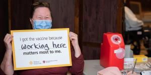 A Hebrew SeniorLife employee sits in a chair, wearing a surgical mask with a sign that says “I got the vaccine because working here matters most to me.” The words “working here” are handwritten.