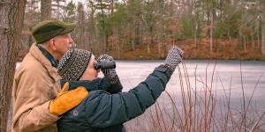 Outdoor winter scene in woods and water with man who has an arm around a woman who is looking through binoculars and pointing.