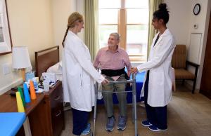 Older man sitting on bed smiling up at two nurses who are readying a walker