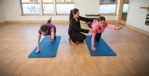 Two women crouching on yoga mats with outstretched arm and leg receiving guidance from a trainer.tw