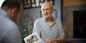 Older man sitting while another man is looing at a photo book