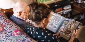 Older man sitting down on a couch looking at a photo album with a woman next to him.