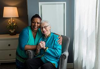 A caretaker hugs an older woman sitting in a chair as they both smile.