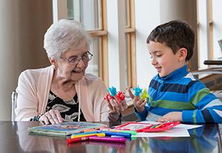 Older woman sits at a table with a young boy as they play with colorful finger puppets.