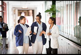 Two female student nurses and a female nurse stand talking in a hallway at Hebrew Rehabilitation Center in Boston. The students are wearing short blue coats and the nurse is wearing a white coat.